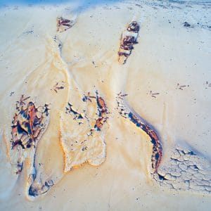 Abstract image of pale sand and sky-blue tones with branching, river-like cracks and rust-colored fragments, forming an organic pattern that resembles aerial desert terrain, dried riverbeds, and wind-shaped erosion.