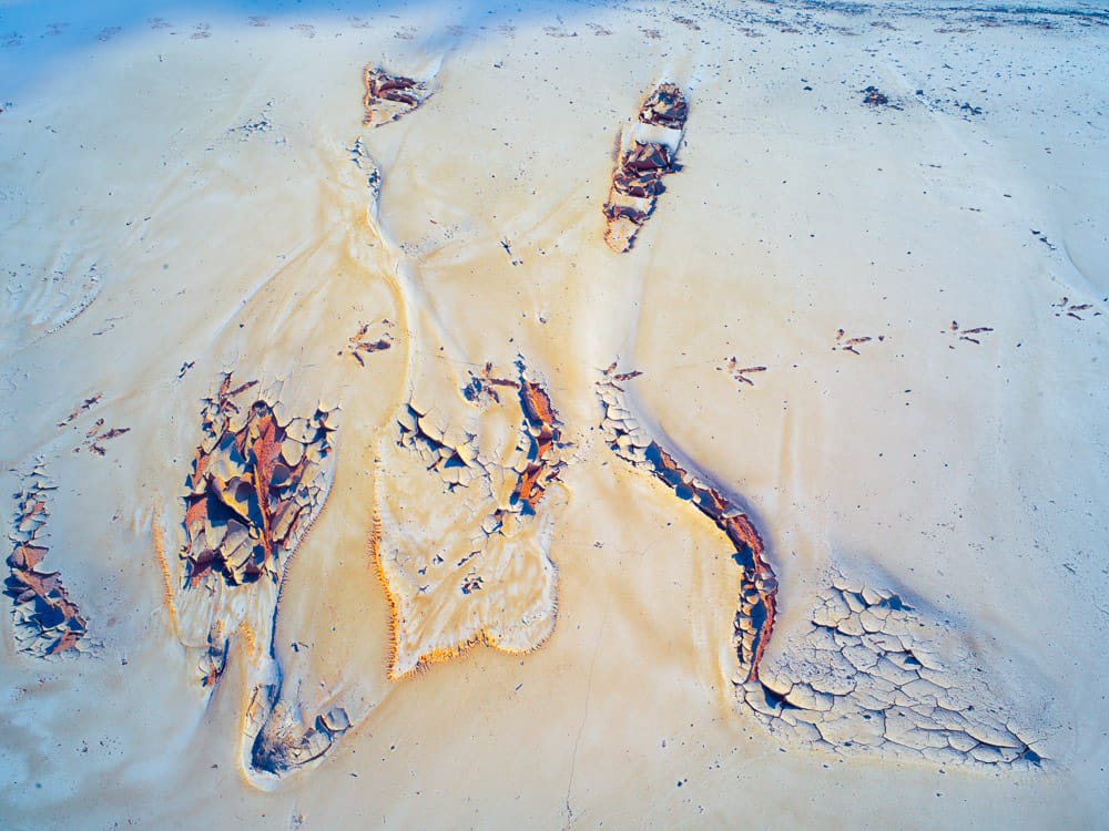 Abstract image of pale sand and sky-blue tones with branching, river-like cracks and rust-colored fragments, forming an organic pattern that resembles aerial desert terrain, dried riverbeds, and wind-shaped erosion.