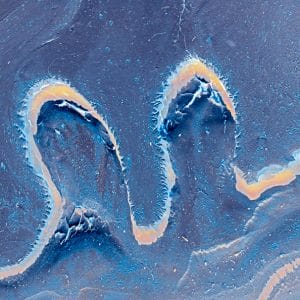 Abstract image of a sinuous, ribbon-like line in pale peach and white cutting through deep blue and slate textures, forming wave-like curves and layered surfaces that suggest fluid motion, erosion, and energy.