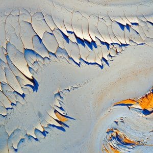 Abstract image of peeling, weathered paint in layered curves, revealing pale blue, deep cobalt shadows, and warm orange-gold textures, forming an organic, flowing pattern reminiscent of eroded stone or wind-carved terrain.