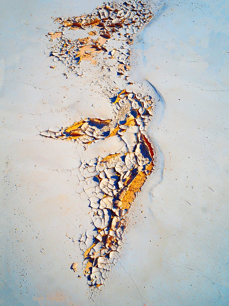 Abstract image of a vertical, river-like fracture of cracked white and ochre textures running through a pale blue surface, with peeling layers and granular details that evoke eroded stone, dried clay, and a weathered geological channel.