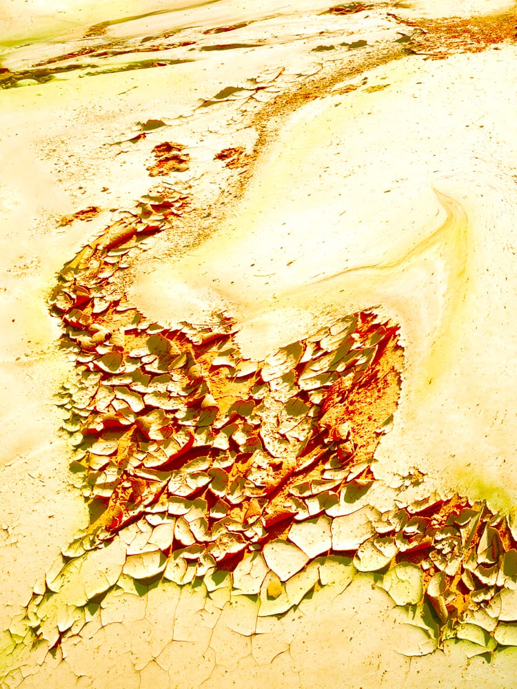 Abstract image of sunlit yellow and cream textures with cracked, peeling layers in red and green, forming a flowing diagonal band that evokes eroded desert stone, wind-worn surfaces, and glowing geological strata.