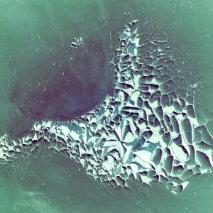 Abstract image of a fractured, crystalline formation in pale white and violet tones emerging from a soft green and teal field, with cracked, tessellated textures that resemble ice floes, mineral shards, or an aerial view of broken terrain.