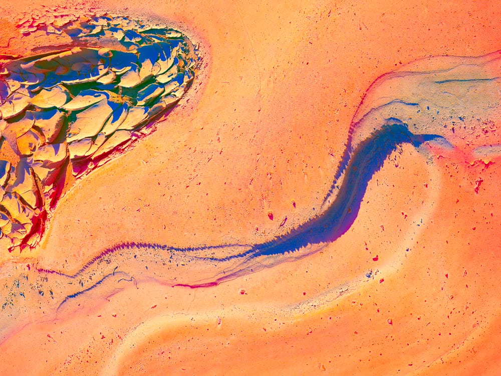 Abstract image of a glowing orange and coral surface crossed by a sinuous blue-violet channel, with cracked, layered textures and fragmented shapes that resemble eroded rock, flowing pigment, and a river cutting through sunlit terrain.