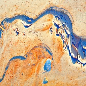 Abstract image of a winding blue ridge cutting through sandy ochre and cream textures, with cracked, peeling layers and scattered blue forms that resemble eroded stone, wind-carved dunes, and geological strata.